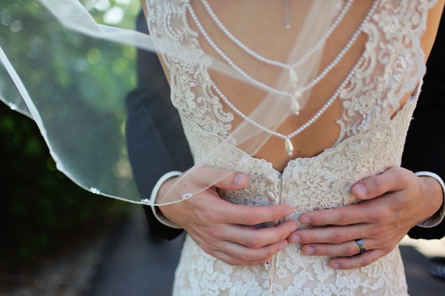 Ruce ženicha objímající nevěstu stojící zády k objektivu-detail šatů-perly